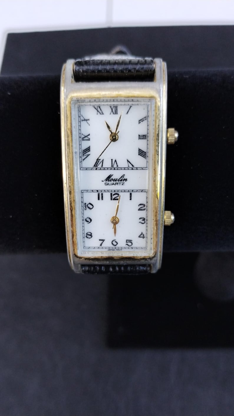 Pu&ograve; includere: Un orologio al quarzo rettangolare con quadrante bianco e numeri romani neri. L'orologio ha una cornice color oro e un cinturino in pelle nera. Il quadrante &egrave; diviso in due sezioni, con il marchio "Moulin Quartz" stampato sulla sezione superiore.