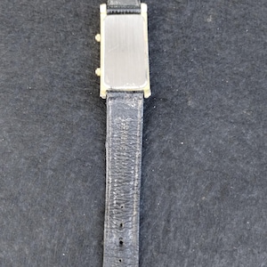 Pu&ograve; includere: Un orologio da polso rettangolare color oro con quadrante argentato e cinturino in pelle nera. L'orologio ha una fibbia dorata ed &egrave; esposto su uno sfondo scuro. Il cinturino mostra segni di usura.