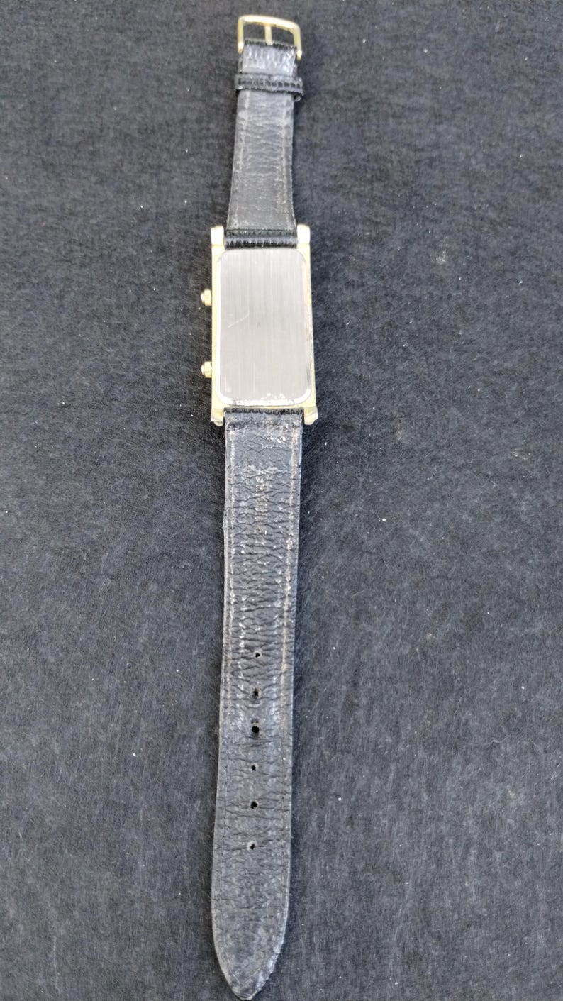 Pu&ograve; includere: Un orologio da polso rettangolare color oro con quadrante argentato e cinturino in pelle nera. L'orologio ha una fibbia dorata ed &egrave; esposto su uno sfondo scuro. Il cinturino mostra segni di usura.