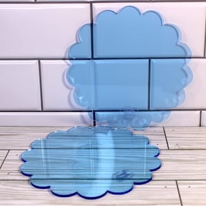 May include: Two transparent blue, scalloped-edge coasters. The coasters are made of a clear material and are placed on a light wood-grain surface. The background features white tiles. The coasters have a cloud-like shape.