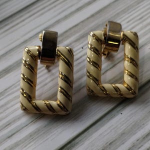 May include: A pair of square earrings with a cream-colored enamel finish and gold-tone accents. The square frames have a twisted rope design. The earrings have a gold-tone post and a rectangular top.