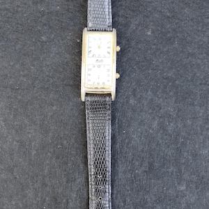 Pu&ograve; includere: Un orologio da polso rettangolare con cassa color oro e cinturino in pelle nera. Il quadrante dell'orologio &egrave; bianco con numeri e lancette nere. Il cinturino ha un motivo strutturato, simile alla pelle di rettile, e una fibbia dorata.