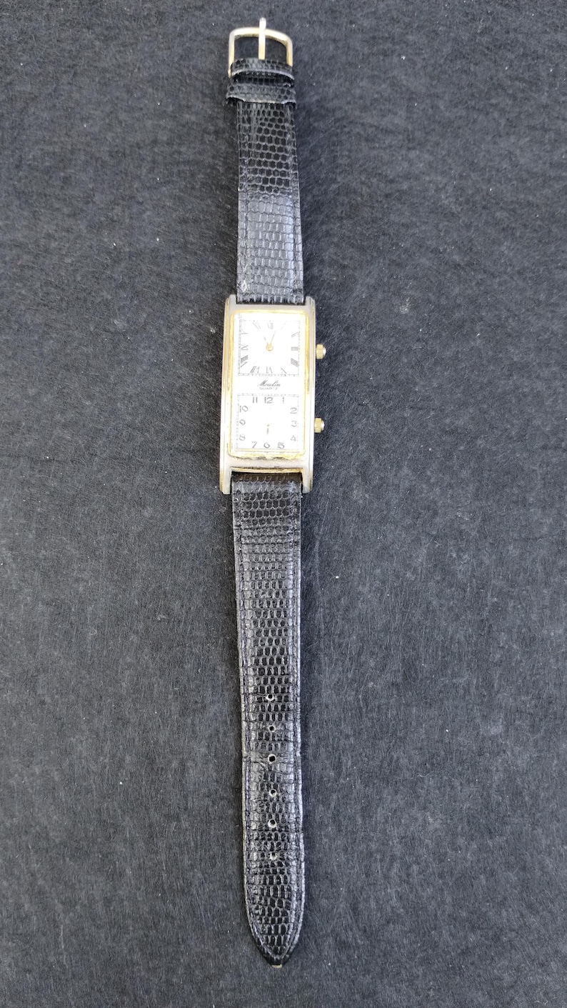 Pu&ograve; includere: Un orologio da polso rettangolare con cassa color oro e cinturino in pelle nera. Il quadrante dell'orologio &egrave; bianco con numeri e lancette nere. Il cinturino ha un motivo strutturato, simile alla pelle di rettile, e una fibbia dorata.