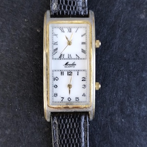 Könnte beinhalten: Eine rechteckige Armbanduhr mit einem silber- und goldfarbenen Gehäuse und einem schwarzen Lederarmband. Das Zifferblatt der Uhr ist in zwei Abschnitte unterteilt, die jeweils eine separate Zeit anzeigen. Der obere Abschnitt hat römische Ziffern, und der untere Abschnitt hat arabische Ziffern. Die Uhr zeigt "Moulin Quartz".