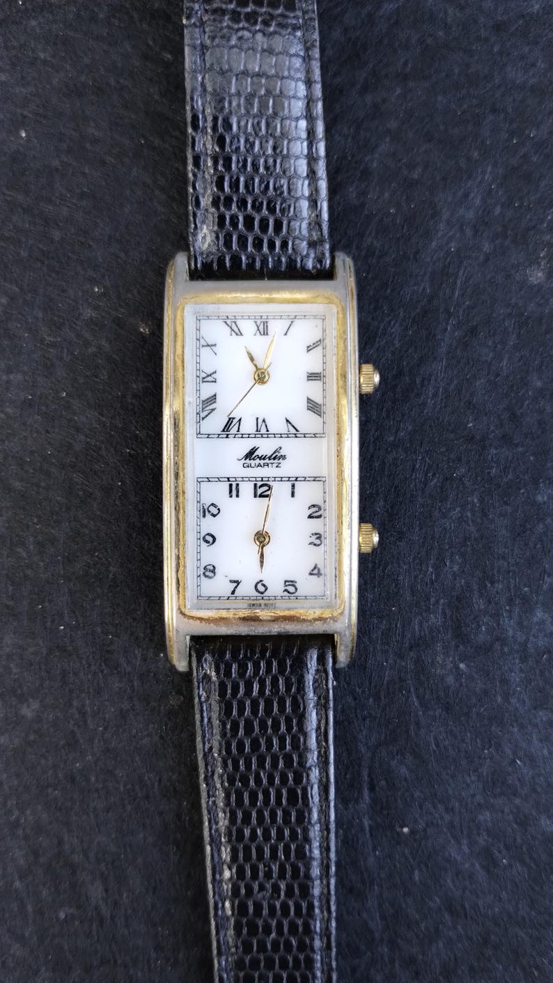 Pu&ograve; includere: Un orologio da polso rettangolare con cassa argentata e dorata e cinturino in pelle nera. Il quadrante dell'orologio &egrave; diviso in due sezioni, ciascuna delle quali mostra un'ora diversa. La sezione superiore ha numeri romani e la sezione inferiore ha numeri arabi. L'orologio indica "Moulin Quartz".