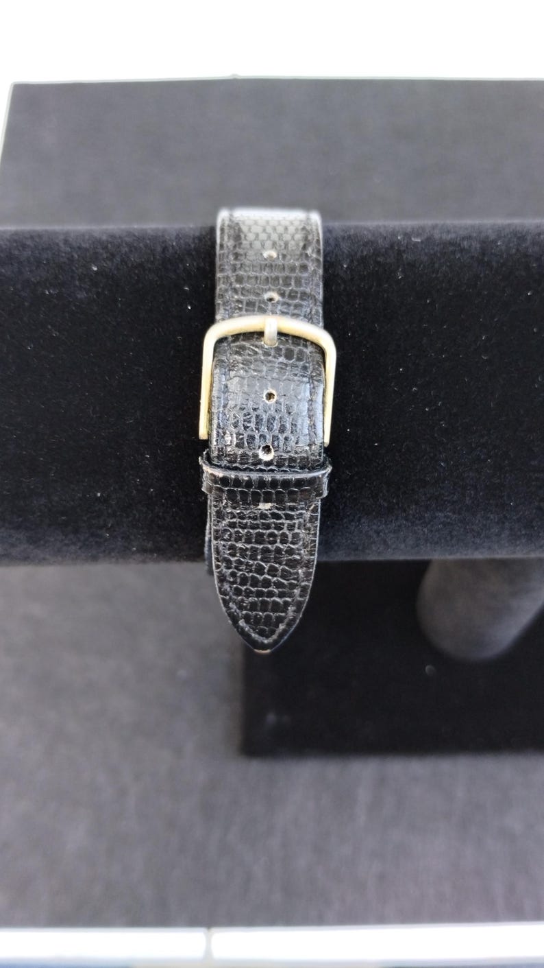 Pu&ograve; includere: Primo piano di un cinturino nero per orologio con un motivo strutturato simile a quello di un rettile. Il cinturino presenta una fibbia color oro ed &egrave; ambientato su uno sfondo scuro, evidenziando i dettagli della pelle e dell'hardware.