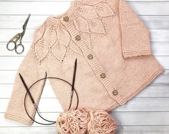 May include: A pink knitted cardigan with a leaf pattern and wooden buttons. The cardigan is laid flat on a white wooden surface with two balls of pink yarn and knitting needles.
