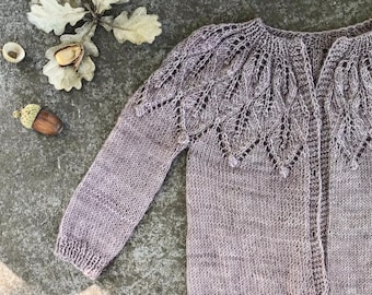 May include: A light brown knitted cardigan with a leaf-like pattern around the neckline and shoulders. The long-sleeved sweater has a button-up front and is displayed on a textured surface with acorns and leaves.