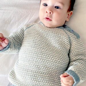 May include: A baby wearing a light blue and white knit sweater with blue trim. The sweater has a textured pattern and button details on the shoulder. The baby is lying on a white surface.