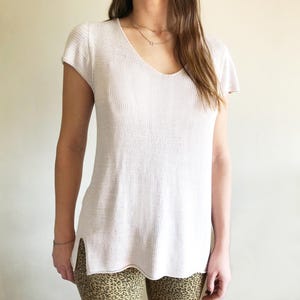 May include: A light pink knit top with a V-neck and short sleeves. The top has a loose fit and side slits at the hem. The fabric appears to be a soft, breathable material. The top is a neutral color, suitable for various outfits.