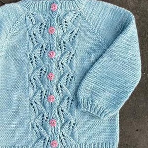 Puede incluir: Cárdigan de punto azul claro con un diseño de punto trenzado en la parte delantera y las mangas. El cárdigan tiene un cuello redondo y se abrocha con seis botones rosas. Las mangas son largas y tienen puños.