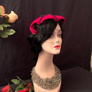 May include: A mannequin wearing a pink velvet hat with a black veil and a silver beaded necklace.