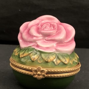 Peut inclure: Une boîte à bijoux en porcelaine en forme de rose. La rose est rose avec des feuilles vertes et une bordure dorée. La boîte a un couvercle à charnière.