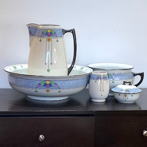May include: A vintage bathroom set with an Art Deco design. The set includes a pitcher, a bowl, a chamber pot, and a lidded jar. The pieces are white with blue, green, and yellow geometric patterns.