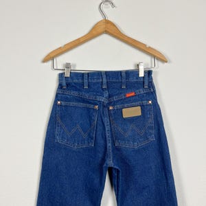 Op de afbeelding: Een paar blauwe denim jeans met een bruin leren patch op de achterzak met de tekst "Wrangler".