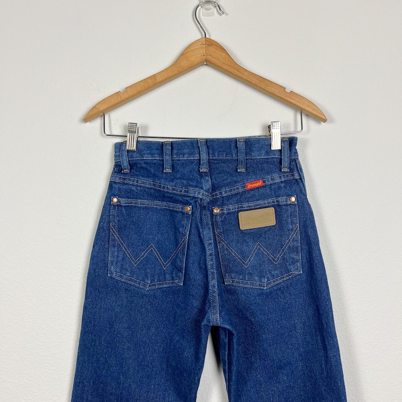 Op de afbeelding: Een paar blauwe denim jeans met een bruin leren patch op de achterzak met de tekst "Wrangler".