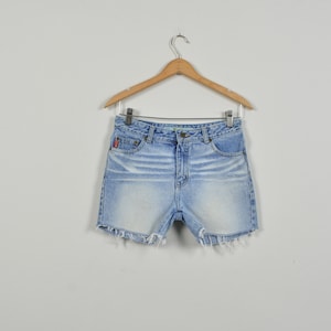 May include: Light blue denim shorts with a faded wash and frayed hem, hanging on a wooden hanger. The shorts have a classic five-pocket design and a red logo patch on the back pocket.
