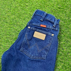 Op de afbeelding: Een donkerblauwe spijkerbroek met een bruin leren patch met de tekst "Wrangler" op de achterzak.