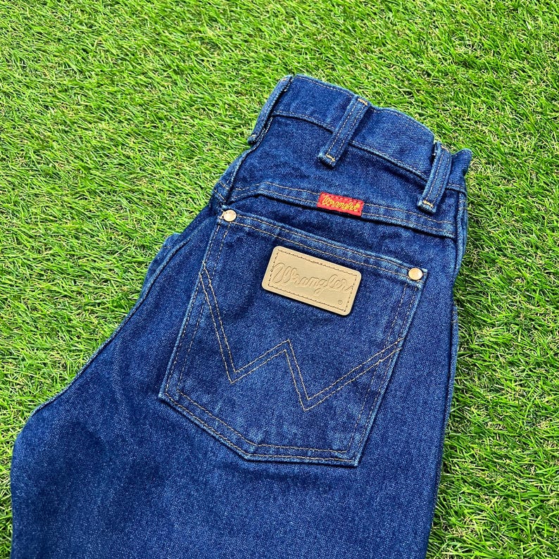 Op de afbeelding: Een donkerblauwe spijkerbroek met een bruin leren patch met de tekst "Wrangler" op de achterzak.