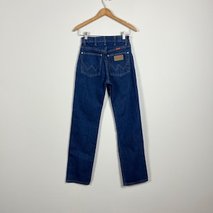Op de afbeelding: Een paar donkerblauwe spijkerbroeken met een bruin leren patch op de achterzak met de tekst "Wrangler".