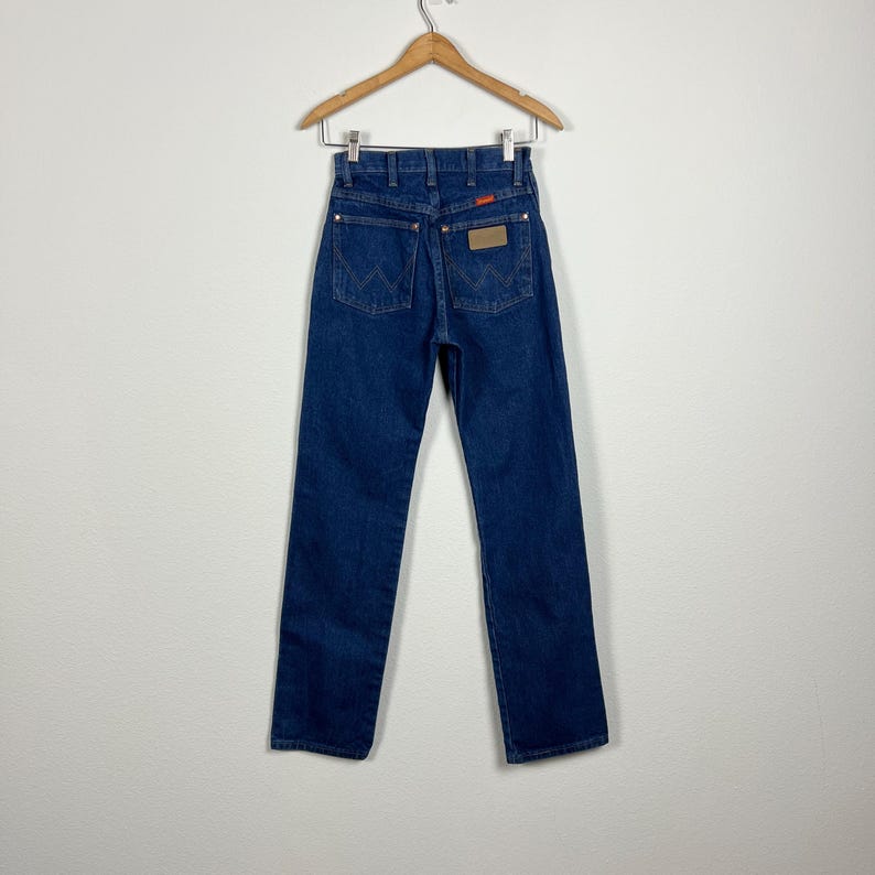 Op de afbeelding: Een paar donkerblauwe spijkerbroeken met een bruin leren patch op de achterzak met de tekst "Wrangler".
