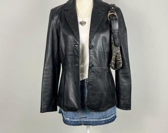 May include: A black leather blazer with a button-up closure and a silver heart pendant necklace. The blazer is worn over a white button-up shirt and a denim skirt.