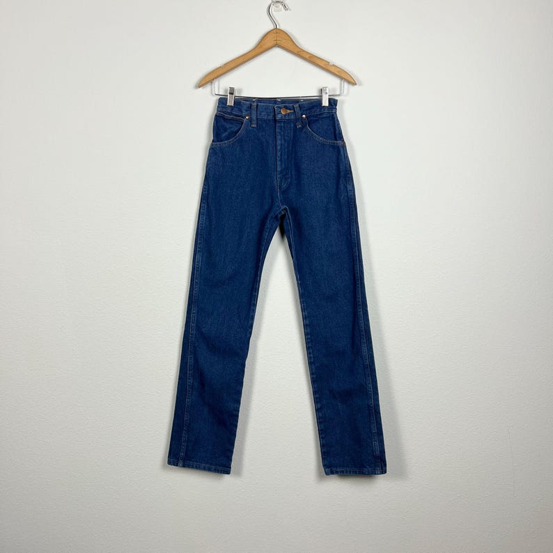 Op de afbeelding: Een donkerblauwe spijkerbroek met rechte pijpen die aan een houten hanger hangt. De broek heeft een hoge taille.