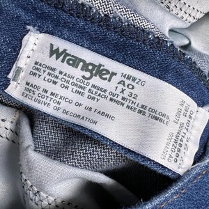 Op de afbeelding: Een wit Wrangler-jeansverzorgingsetiket met blauwe tekst. Het etiket zegt "Machinewas koud binnenstebuiten met gelijke kleuren, alleen niet-chloorbleekmiddel indien nodig, drogen in de droger op lage temperatuur of aan de lijn drogen. Gemaakt in Mexico van Amerikaanse stof, 100% katoen, exclusief decoratie. 14MWZG 1 X 32"