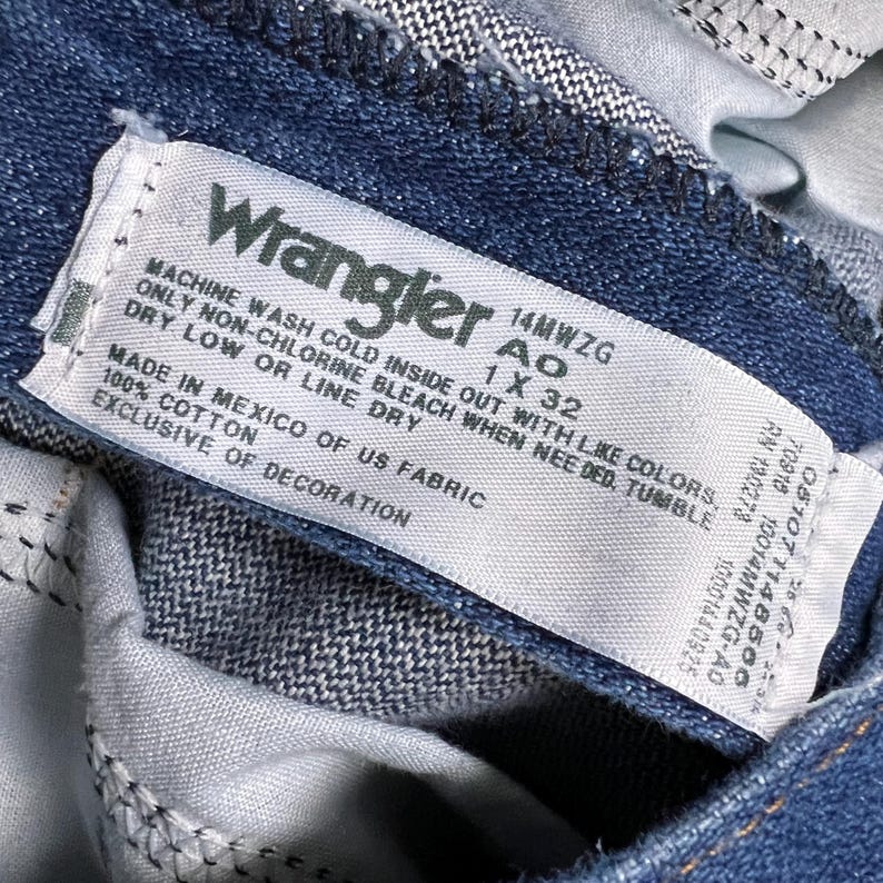 Op de afbeelding: Een wit Wrangler-jeansverzorgingsetiket met blauwe tekst. Het etiket zegt "Machinewas koud binnenstebuiten met gelijke kleuren, alleen niet-chloorbleekmiddel indien nodig, drogen in de droger op lage temperatuur of aan de lijn drogen. Gemaakt in Mexico van Amerikaanse stof, 100% katoen, exclusief decoratie. 14MWZG 1 X 32"