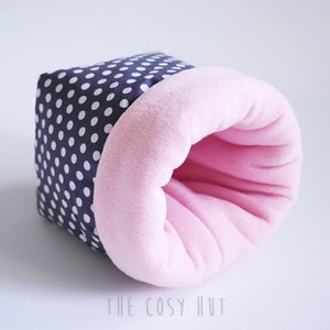 Könnte beinhalten: Ein rosa-weiß gepunktetes Stofftierbett mit rosa Fleece-Futter. Das Bett ist zylinderförmig mit einem Loch in der Mitte.