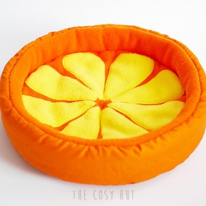 Peut inclure: Lit de compagnie en forme d'orange avec un intérieur en peluche jaune. Le lit est fait de tissu orange et a un design cousu qui ressemble à une tranche d'orange.