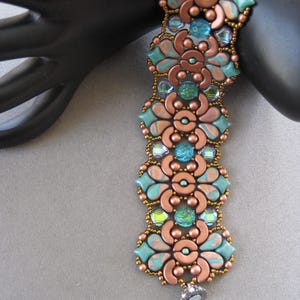 May include: A beaded bracelet featuring a floral design with copper-colored metal and turquoise and blue beads. The bracelet has a magnetic clasp and is displayed on a black mannequin hand. The beads are arranged in a repeating pattern.