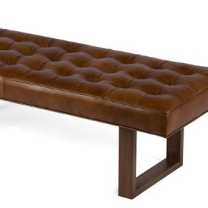 May include: A brown leather bench with a tufted top and wooden legs. The bench has a rectangular shape and is supported by four legs.