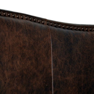 May include: Close-up of a section of a brown leather chair back. The leather has a distressed appearance with visible grain and texture. The top edge is adorned with evenly spaced, dark metal studs.