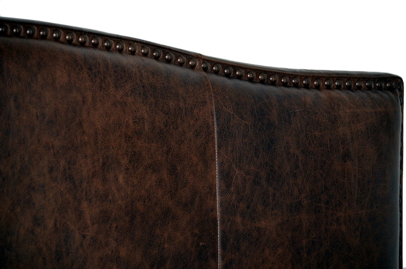 May include: Close-up of a section of a brown leather chair back. The leather has a distressed appearance with visible grain and texture. The top edge is adorned with evenly spaced, dark metal studs.