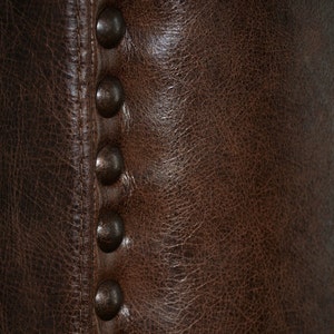 May include: Close-up of a section of a brown leather chair. The leather has a textured appearance with visible grain and subtle variations in color. A row of six dark, rounded metal studs is visible along the seam.
