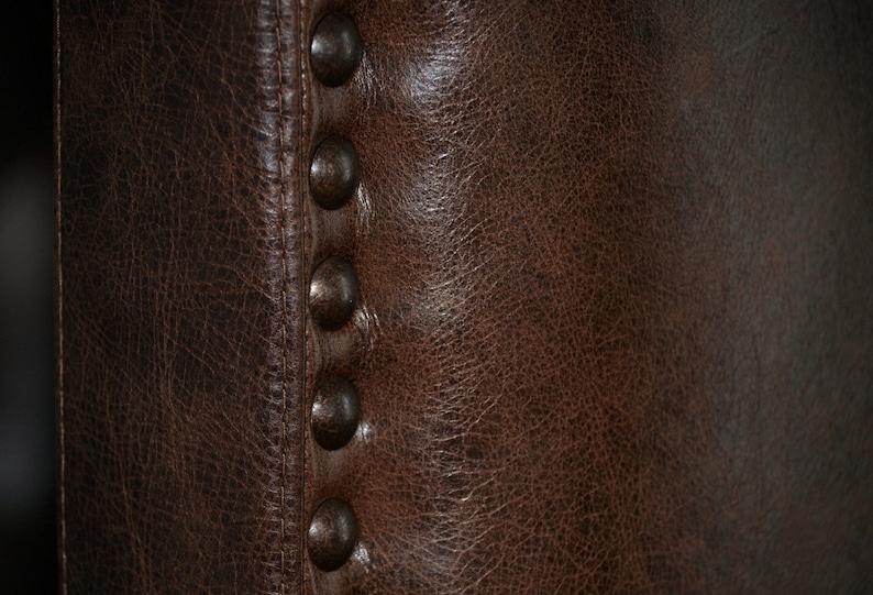 May include: Close-up of a section of a brown leather chair. The leather has a textured appearance with visible grain and subtle variations in color. A row of six dark, rounded metal studs is visible along the seam.