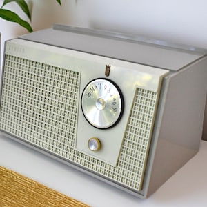 May include: A vintage tabletop radio with a light gray plastic casing and a cream-colored front speaker grille. The radio features a circular silver tuning dial with black numbers and a small gold-toned knob positioned directly below the dial.