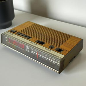 May include: A General Electric digital alarm clock radio with a wood grain top panel, a black speaker grille, and a front-facing display featuring a red digital time readout, an analogue AM/FM frequency tuner, and the model number 7-4924 printed.