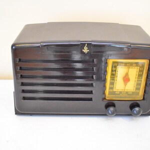 May include: A vintage dark brown radio with a yellow dial and two black knobs. The radio has horizontal vent-like lines on the front and a small gold emblem above the dial. The radio is sitting on a white surface.