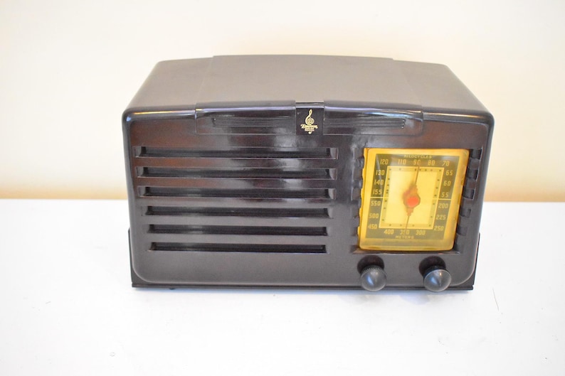 May include: A vintage dark brown radio with a yellow dial and two black knobs. The radio has horizontal vent-like lines on the front and a small gold emblem above the dial. The radio is sitting on a white surface.