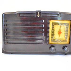 May include: A vintage dark brown radio with a rectangular design. The radio features a speaker grill, a yellowed dial, and two black knobs. A gold emblem is at the top. The radio is likely from the mid-20th century.