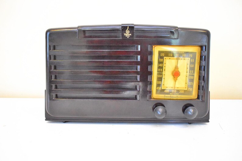 May include: A vintage dark brown radio with a rectangular design. The radio features a speaker grill, a yellowed dial, and two black knobs. A gold emblem is at the top. The radio is likely from the mid-20th century.