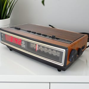 以下が含まれることがあります： 木目調仕上げのビンテージGeneral Electricデジタル目覚まし時計。 時計は赤い数字で時間を表示し、ラジオダイヤルが付いています。 時計は白い表面にあります。
