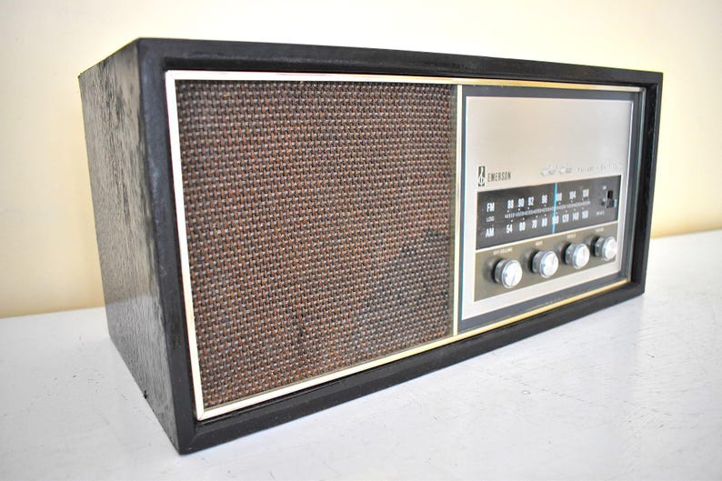 May include: A vintage Emerson AM/FM radio with a woodgrain cabinet and a black speaker grille. The radio has a silver face with a blue tuning dial and silver knobs.