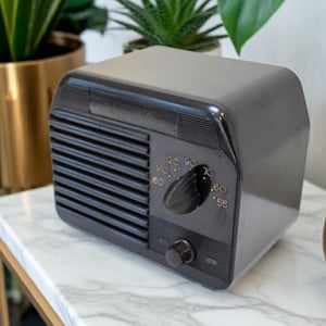 May include: A retro black radio featuring a tuning dial and speaker grill. The radio is placed on a marble-effect surface, with gold decorative items and plants in the background. The dial displays numbers from 55 to 140.