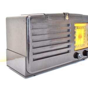 May include: A vintage dark brown radio with a glowing yellow dial. The radio has horizontal vent-like lines and two black knobs. A gold emblem is on the top. The radio is set against a white background.