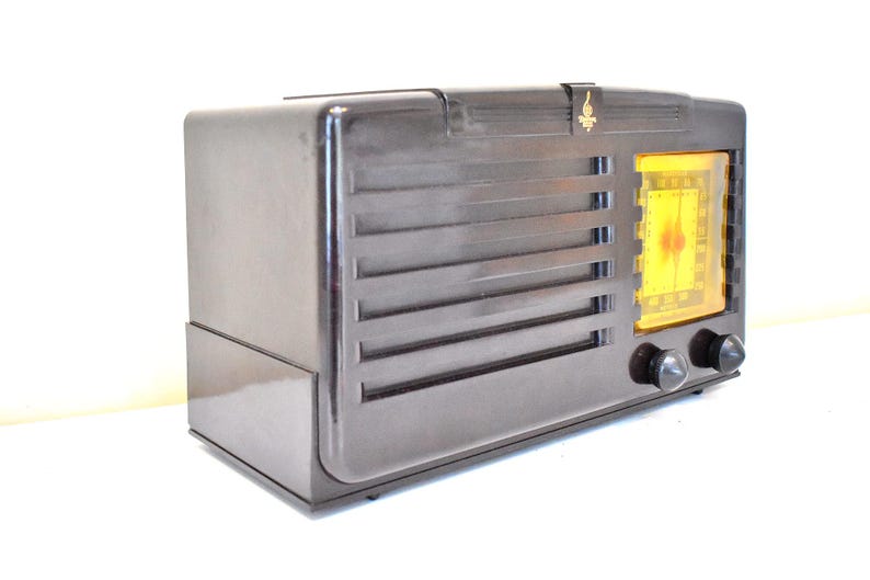 May include: A vintage dark brown radio with a glowing yellow dial. The radio has horizontal vent-like lines and two black knobs. A gold emblem is on the top. The radio is set against a white background.
