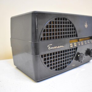 Puede incluir: Una radio Emerson negra vintage con una cubierta de altavoz cuadrada con patr&oacute;n de rejilla. La radio tiene un dial de sintonizaci&oacute;n y dos perillas negras. La palabra "Emerson" est&aacute; impresa en oro en la parte delantera.