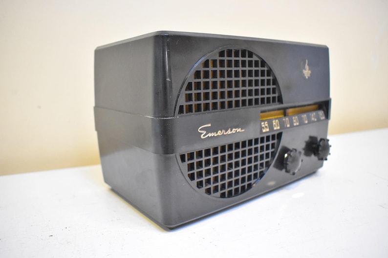 Puede incluir: Una radio Emerson negra vintage con una cubierta de altavoz cuadrada con patr&oacute;n de rejilla. La radio tiene un dial de sintonizaci&oacute;n y dos perillas negras. La palabra "Emerson" est&aacute; impresa en oro en la parte delantera.